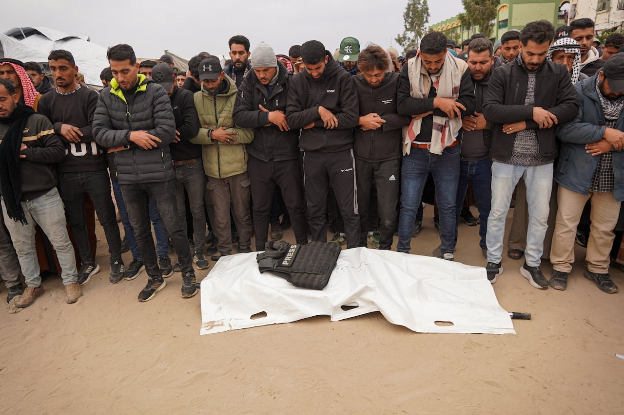 A group of individuals stand beside a covered deceased body with a vest labeled 'Press'.